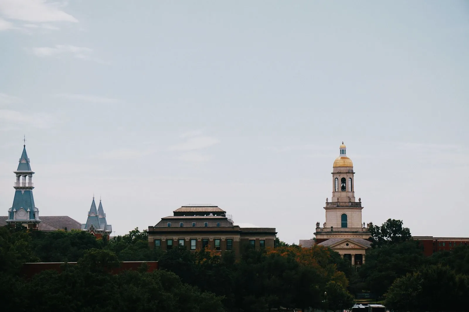 A scenic view featuring historic buildings and towering spires against a clear sky, surrounded by lush trees.